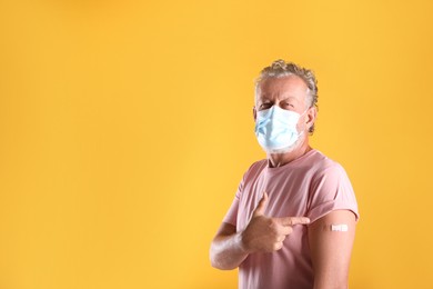 Senior man in protective mask pointing at arm with bandage after vaccination on yellow background. Space for text
