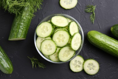 Cut cucumber in glass bowl, fresh vegetables and dill on dark gray textured table, flat lay