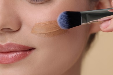Woman applying foundation on face with brush against beige background, closeup