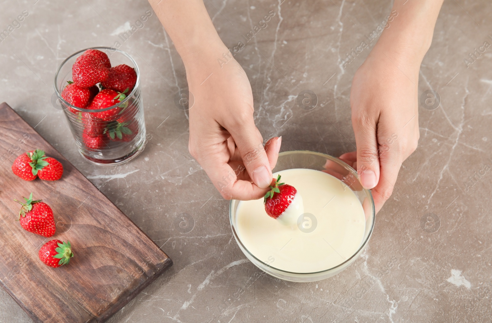 Photo of Woman dipping ripe strawberry into bowl with white melted chocolate on table