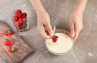 Photo of Woman dipping ripe strawberry into bowl with white melted chocolate on table