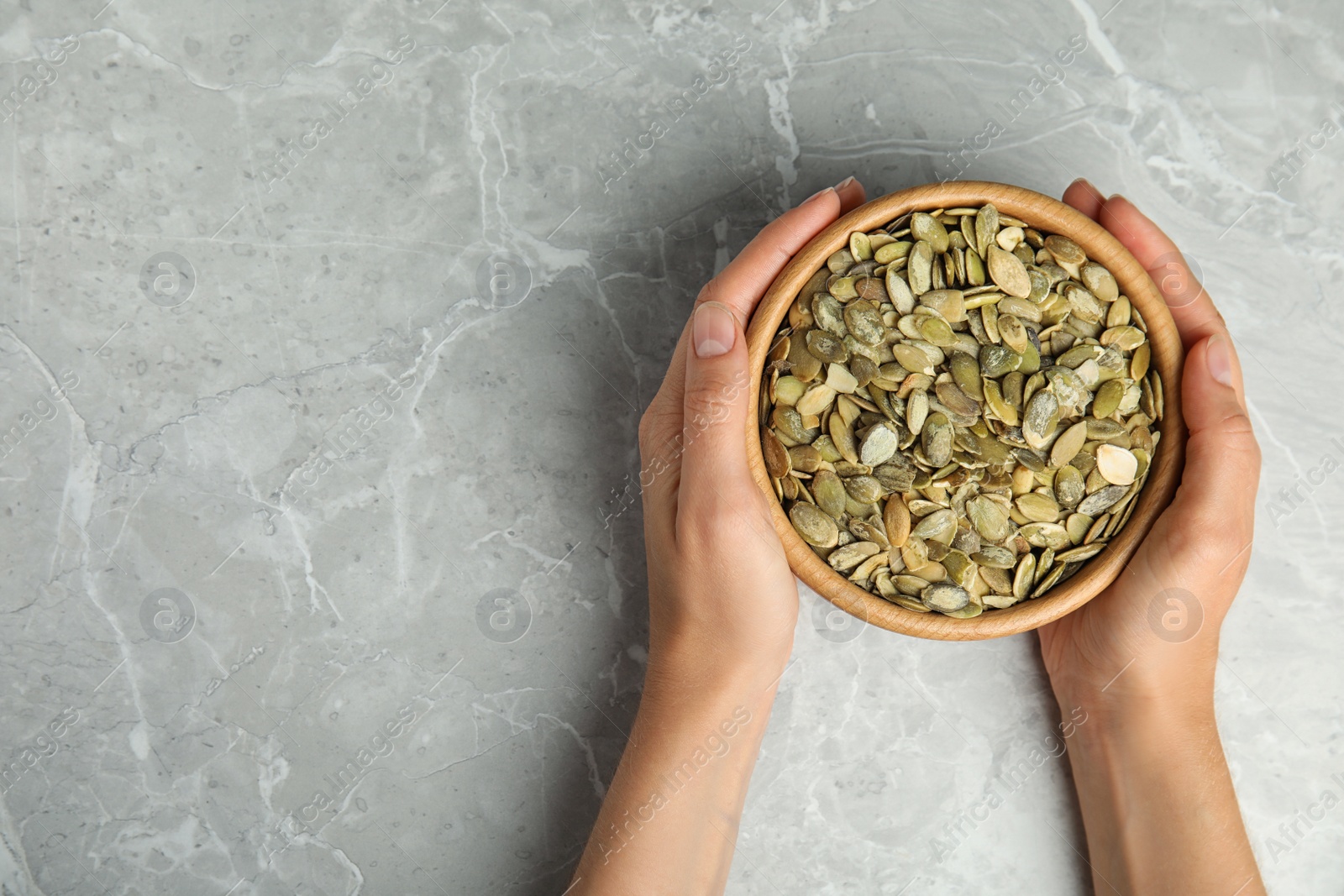 Photo of Young woman with bowl of raw pumpkin seeds at light grey marble table, top view. Space for text