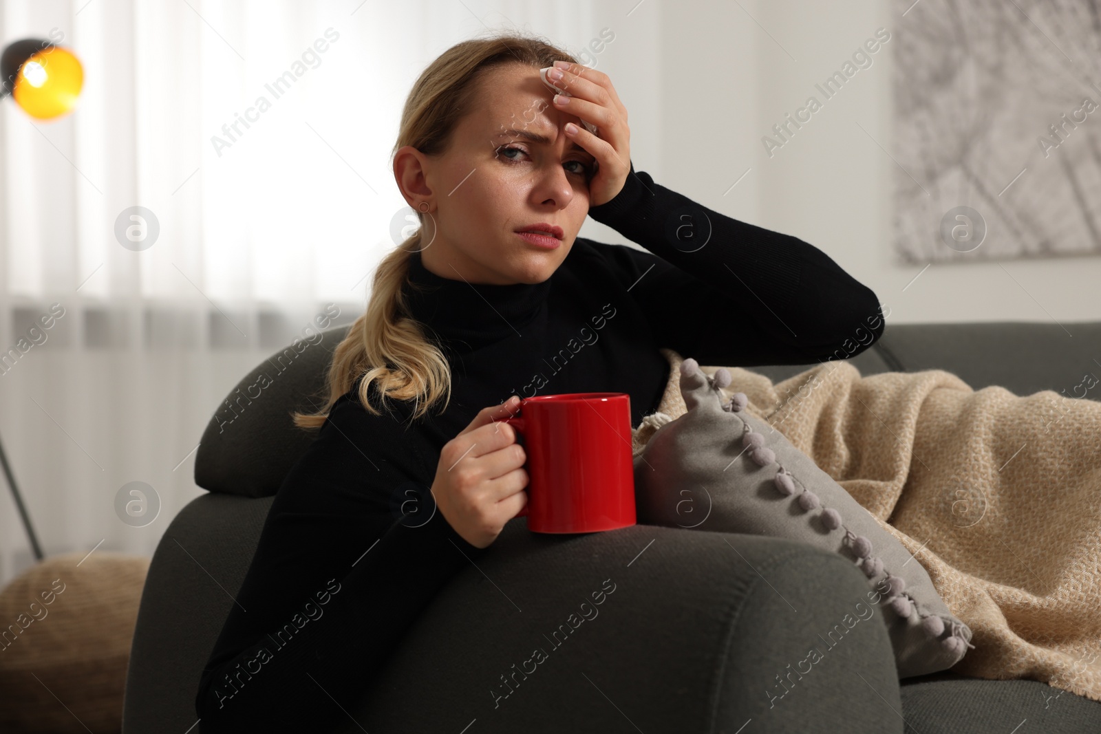 Photo of Sick woman with tissue and cup of drink on sofa at home. Cold symptoms