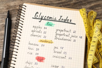 Photo of List with products of low, moderate and high glycemic index in notebook, marker and measuring tape on wooden table, above view
