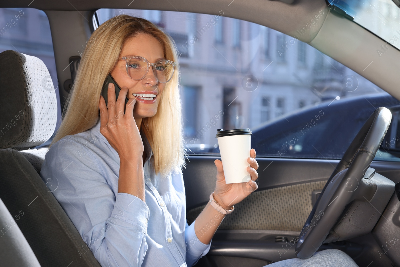 Photo of Coffee to go. Happy woman with paper cup of drink talking on smartphone in car