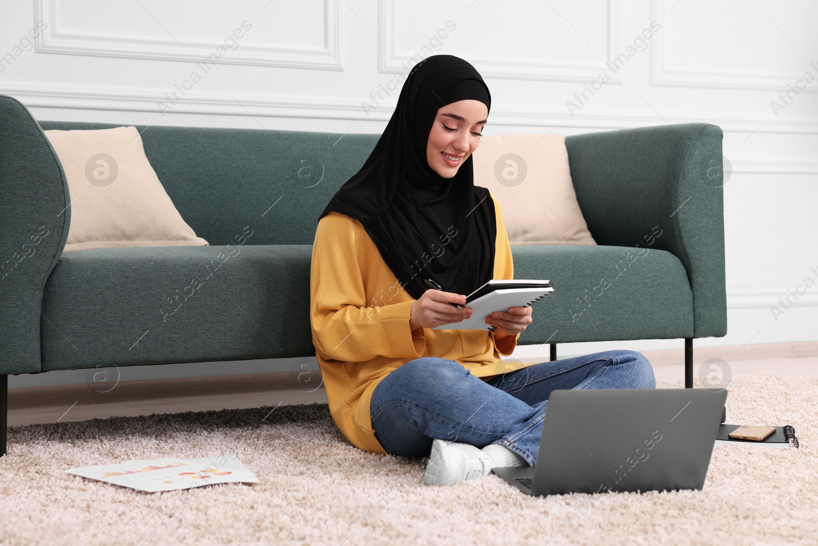 Photo of Muslim woman in hijab using laptop on floor near sofa indoors