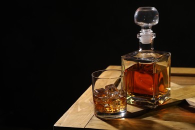 Whiskey with ice cubes in glass and bottle on wooden crate against black background, space for text