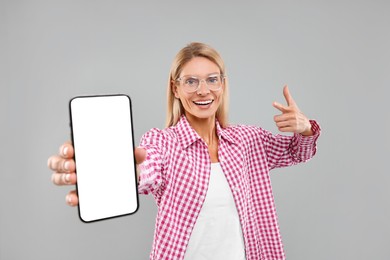 Photo of Happy woman holding smartphone and pointing at blank screen on grey background