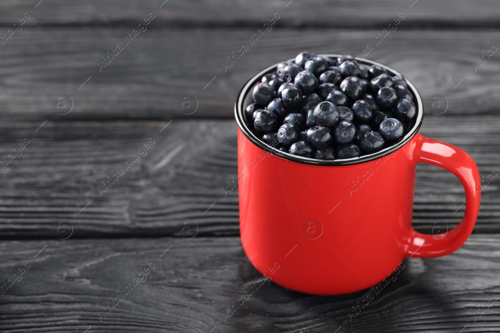 Photo of Red mug of tasty fresh bilberries on black wooden table, closeup. Space for text