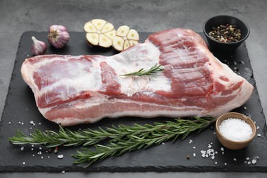 Photo of Piece of raw pork belly, rosemary, garlic and spices on grey table