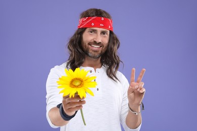 Photo of Stylish hippie man with sunflower showing V-sign on violet background