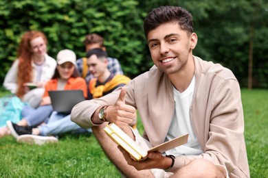 Photo of Students learning together in park. Happy young man with notebook showing thumbs up on green grass, selective focus