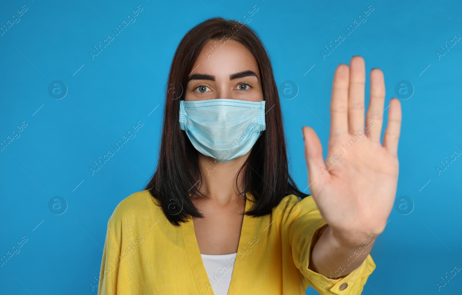 Photo of Young woman in protective mask showing stop gesture on light blue background. Prevent spreading of coronavirus