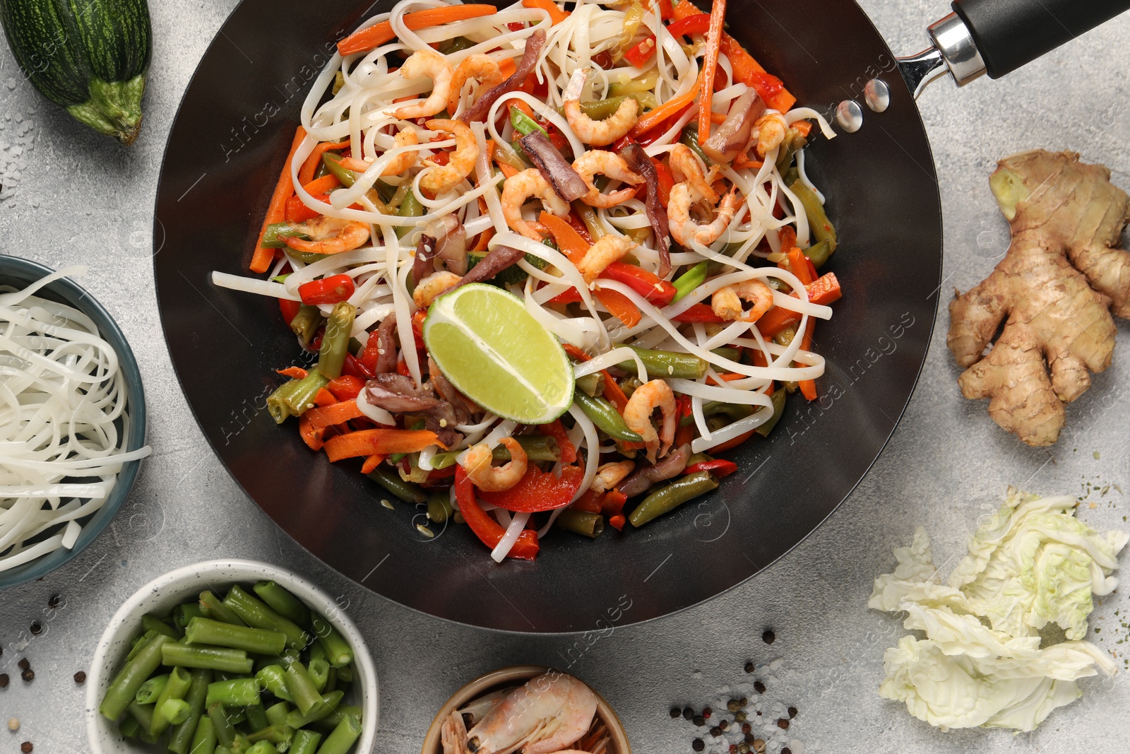 Photo of Shrimp stir fry with noodles and vegetables in wok surrounded by ingredients on grey table, flat lay