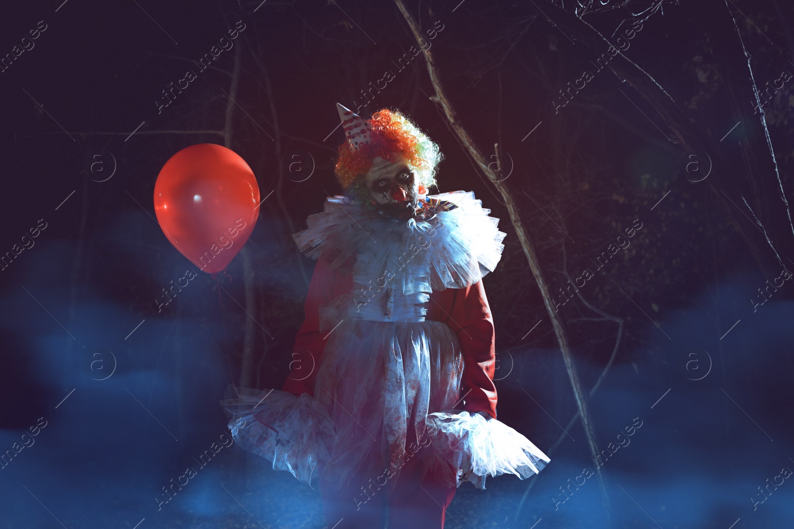 Photo of Terrifying clown with red air balloon outdoors at night. Halloween party costume