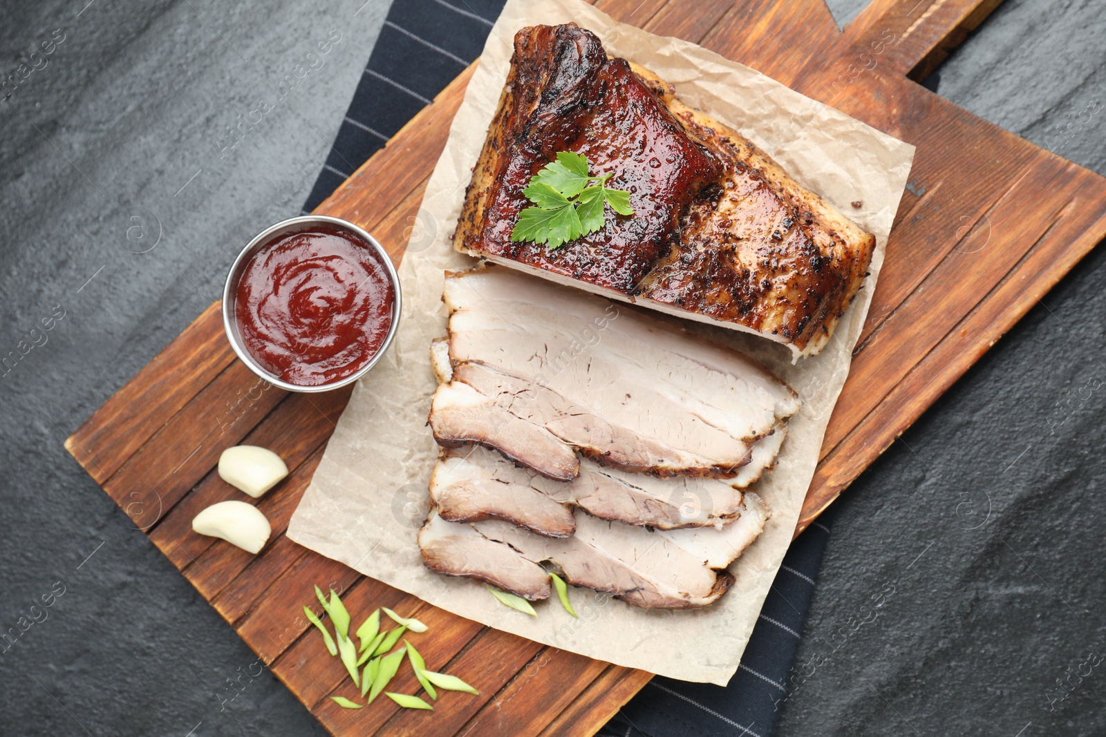 Photo of Pieces of baked pork belly served with sauce and parsley on black textured table, top view