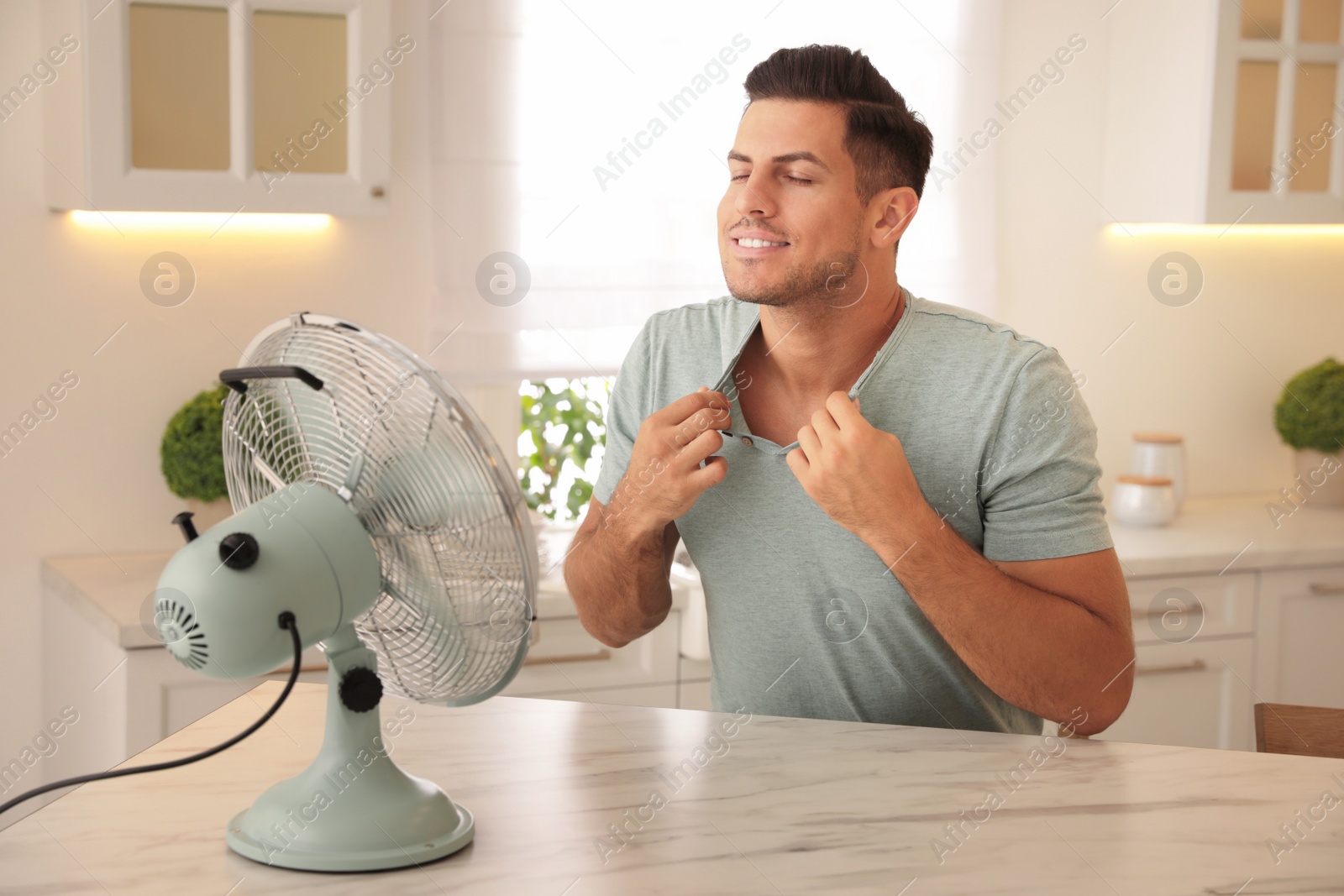 Photo of Man enjoying air flow from fan at table in kitchen. Summer heat