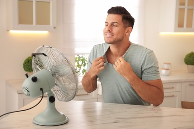 Photo of Man enjoying air flow from fan at table in kitchen. Summer heat