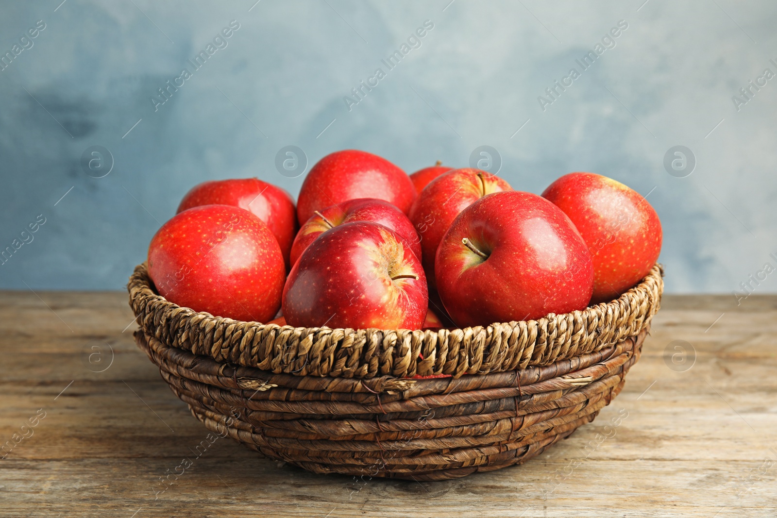 Photo of Wicker bowl with ripe juicy red apples on wooden table against blue background