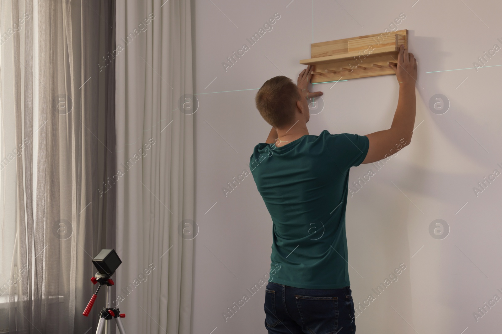 Photo of Man using cross line laser level for hanging wooden shelf on light wall indoors, back view