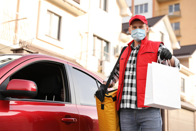 Courier in protective mask with order and thermobag near car outdoors. Food delivery service during coronavirus quarantine