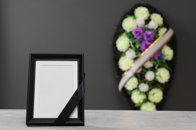 Photo of Photo frame with black ribbon on light table and wreath of flowers near grey wall indoors, space for text. Funeral attributes