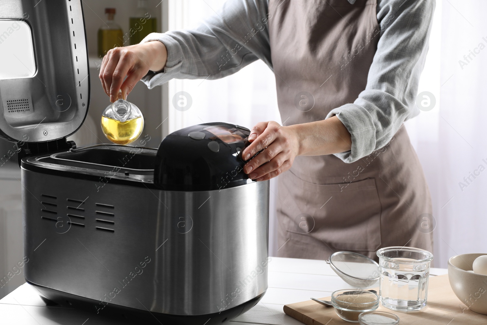 Photo of Woman pouring oil into breadmaker at white wooden table indoors, closeup