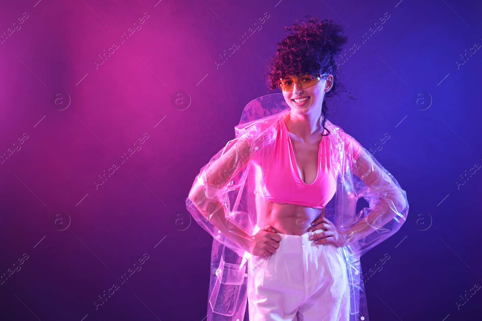 Photo of Beautiful young woman in transparent coat and sunglasses posing on color background in neon lights. Space for text