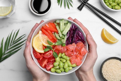 Photo of Woman holding poke bowl with salmon, edamame beans and vegetables at white marble table, top view