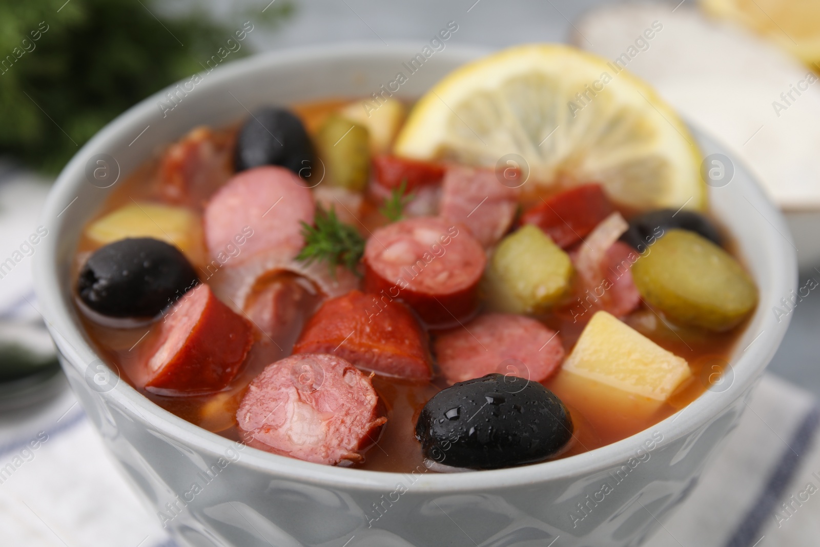 Photo of Meat solyanka soup with thin dry smoked sausages in bowl on table, closeup