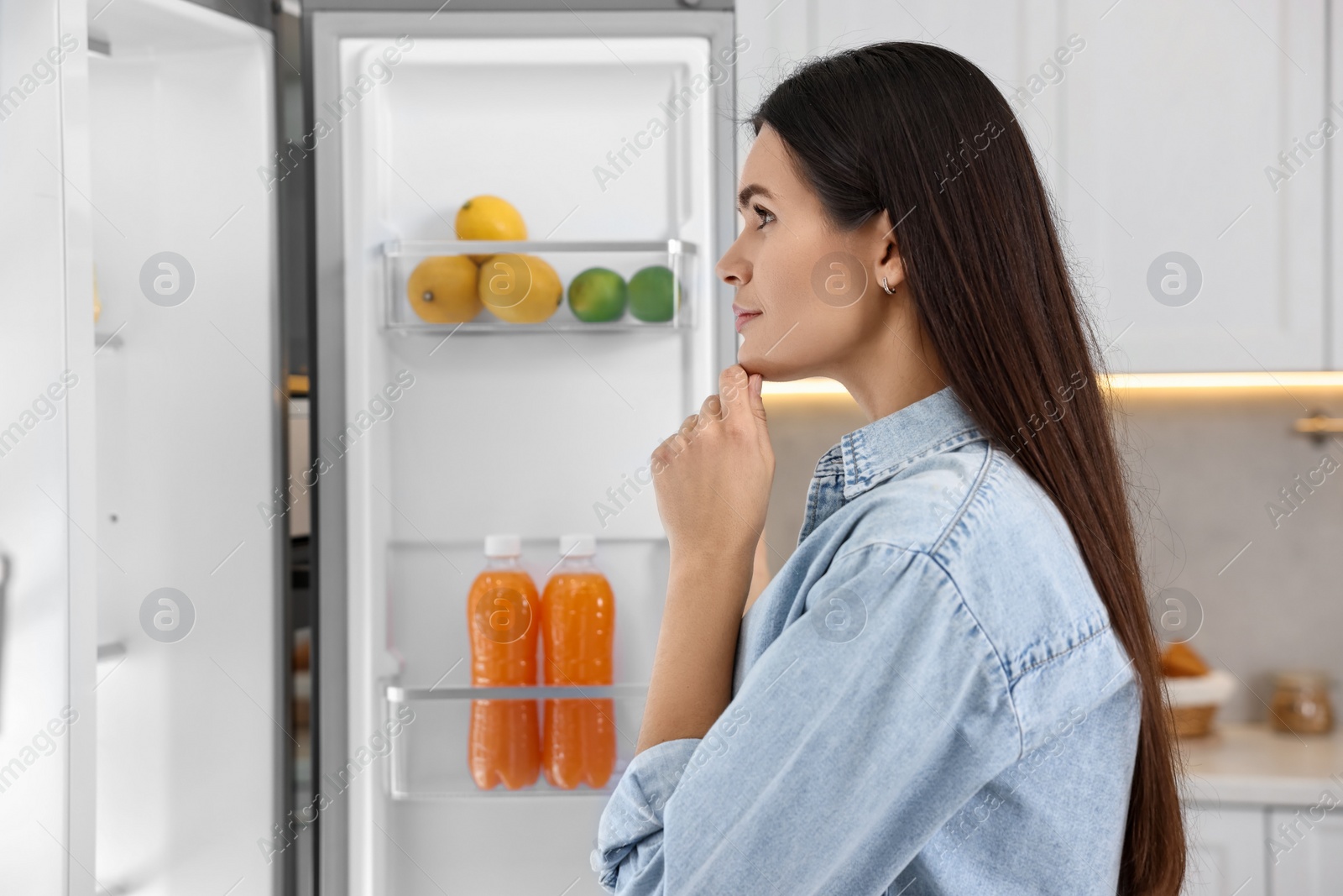 Photo of Thoughtful young woman near modern refrigerator in kitchen