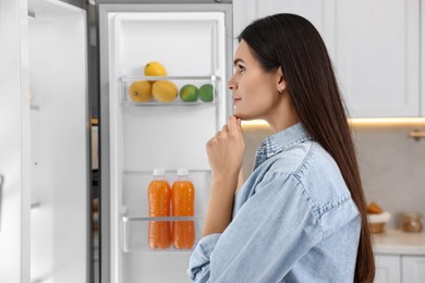 Thoughtful young woman near modern refrigerator in kitchen