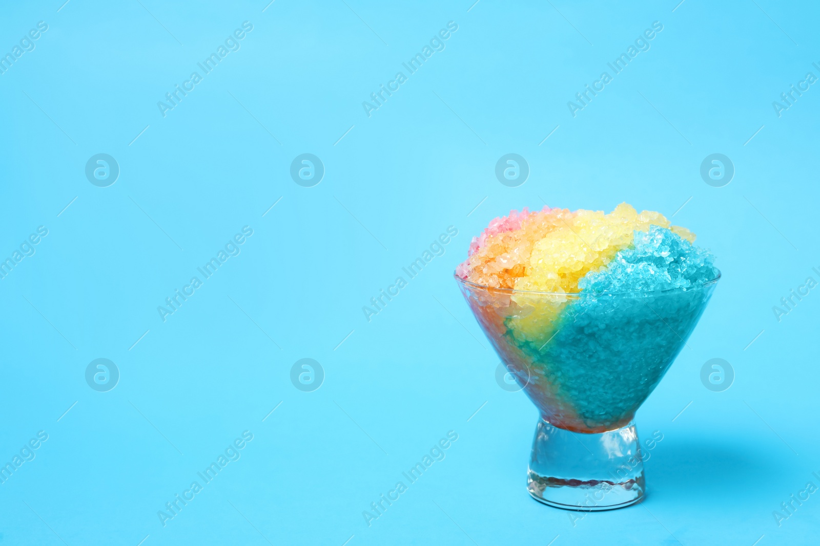 Photo of Rainbow shaving ice in glass dessert bowl on light blue background, space for text