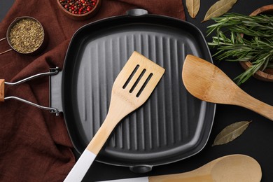 Photo of Wooden kitchen utensils, frying pan and spices on black table, flat lay
