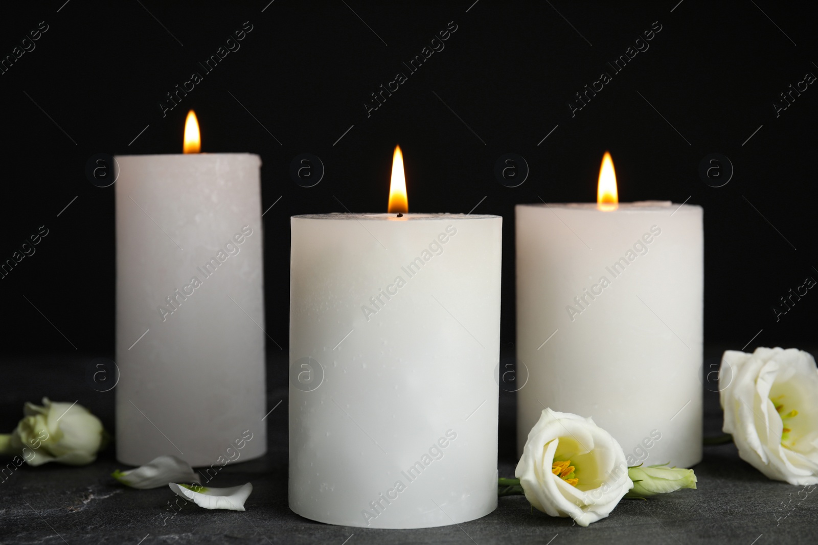 Photo of Burning candles and flowers on grey table against black background