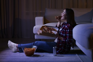 Photo of Young woman eating pizza while watching TV in room at night. Bad habit