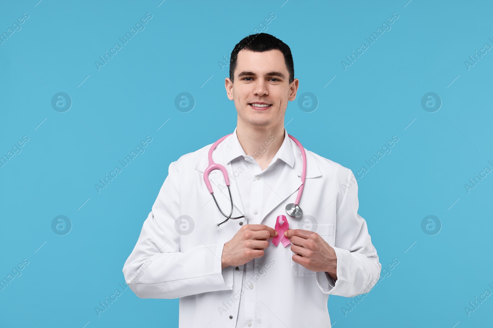 Photo of Portrait of smiling mammologist with pink ribbon and stethoscope on light blue background. Breast cancer awareness