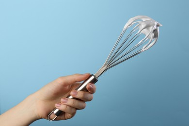 Woman holding whisk with whipped cream on light blue background, closeup