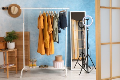 Rack with stylish clothes, mirror and ring lamp near light blue wall indoors