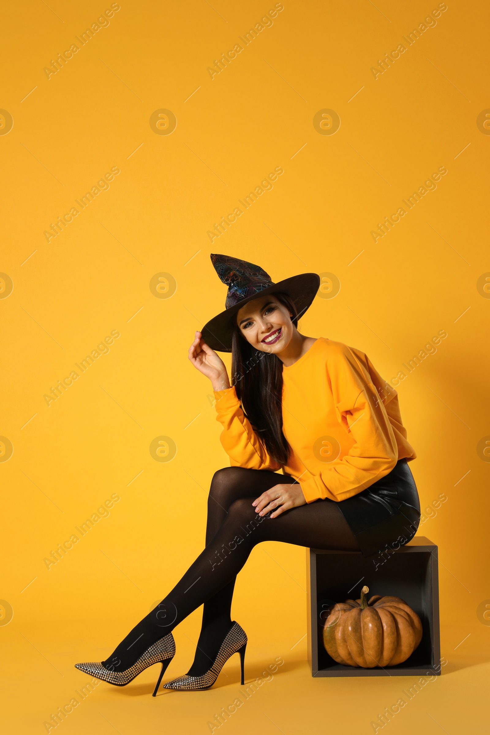 Photo of Beautiful woman wearing witch costume for Halloween party with pumpkin against yellow background