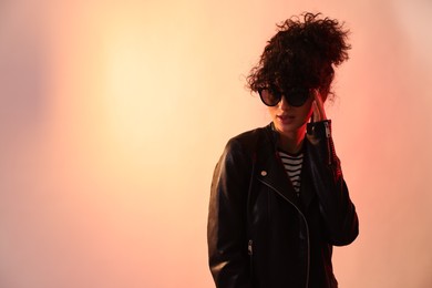 Photo of Beautiful young woman in black leather jacket and sunglasses posing on color background in neon lights. Space for text