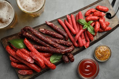 Different thin dry smoked sausages, sauces and glasses of beer on grey table, flat lay