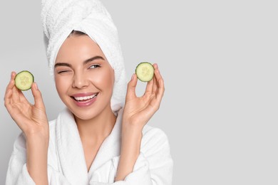 Woman in bathrobe with towel holding pieces of cucumber on light grey background, space for text. Spa treatment