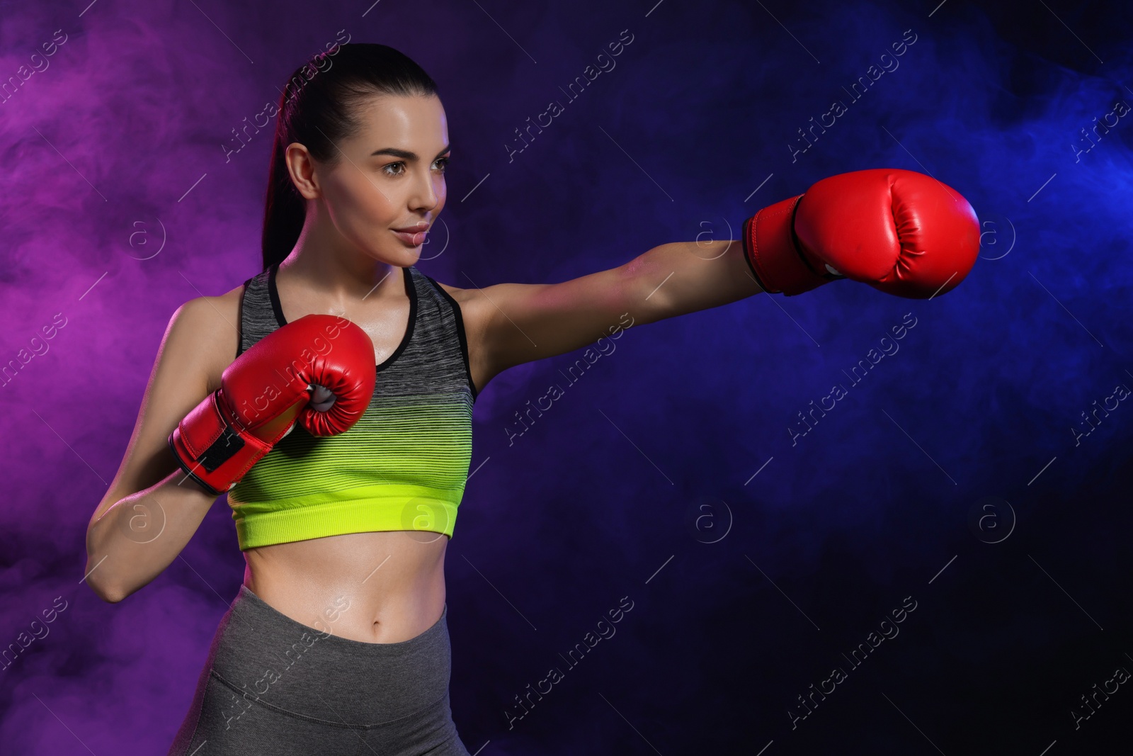 Photo of Portrait of beautiful woman wearing boxing gloves training in color lights and smoke on black background