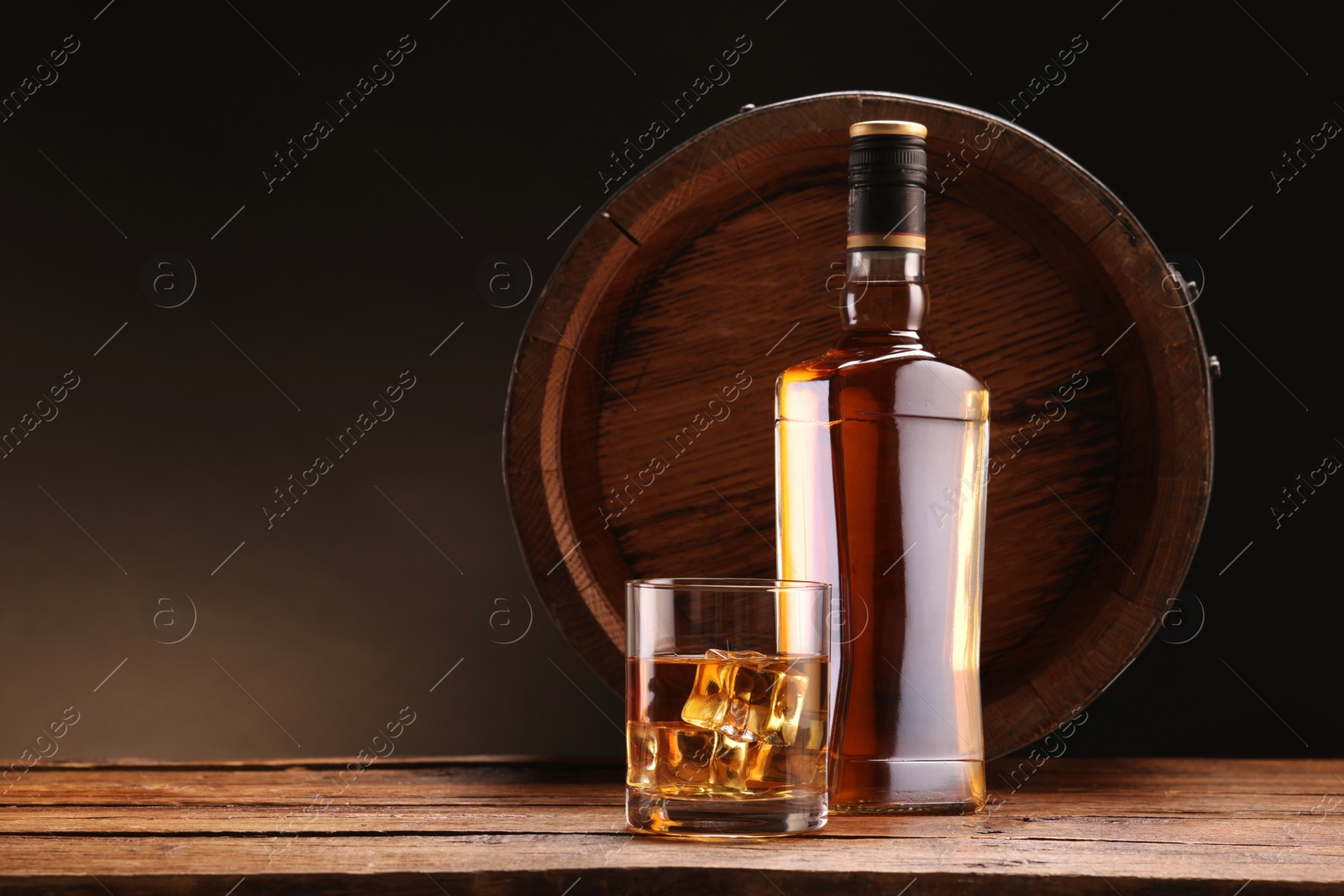 Photo of Whiskey with ice cubes in glass, bottle and barrel on wooden table against black background, space for text