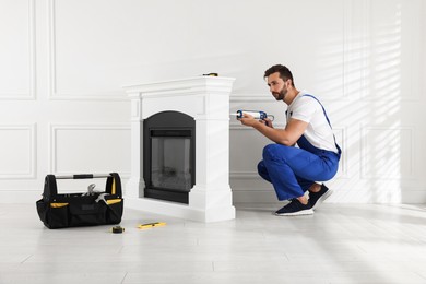 Photo of Professional technician sealing electric fireplace with caulk near white wall in room