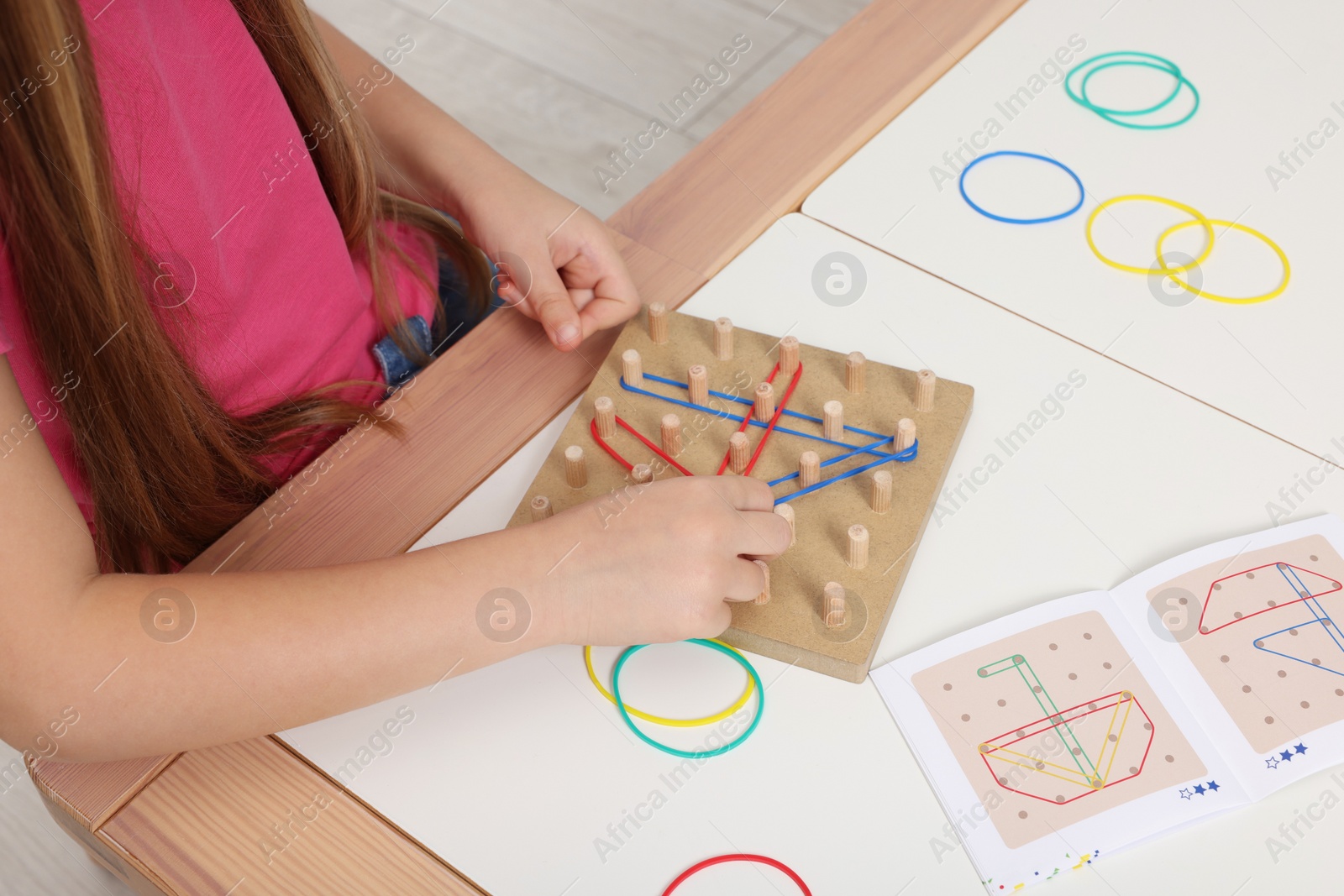 Photo of Motor skills development. Girl playing with geoboard and rubber bands at white table, closeup