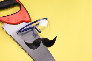 Photo of Man's face made of artificial mustache, safety glasses and hand saw on yellow background, top view. Space for text