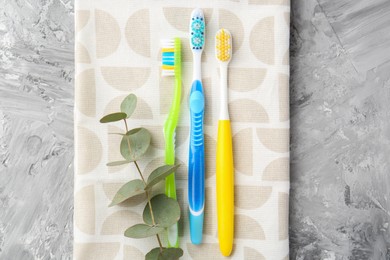 Plastic toothbrushes, towel and eucalyptus branch on grey textured table, top view