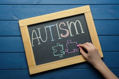 Photo of Woman drawing jigsaw puzzle pieces below word Autism on chalkboard at blue wooden table, top view
